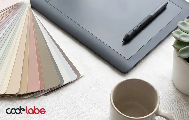 A fan of pastel colour swatches beside a graphics tablet, a stylus, a mug and a small succulent