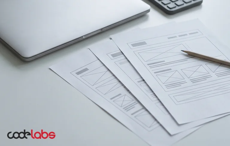 A calculator, a closed laptop and printed website wireframe sheets on a pale grey desk