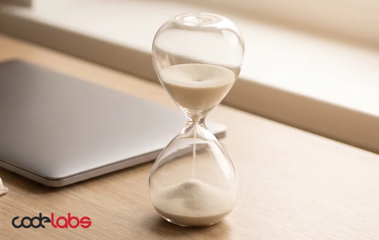An hourglass with sand running through, beside a closed laptop and an open planner on a light oak desk